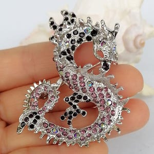 Puede incluir: Broche de drag&oacute;n plateado con pedrer&iacute;a rosa y negra. El dise&ntilde;o del drag&oacute;n presenta escamas detalladas y un cierre de alfiler. Broche decorativo.