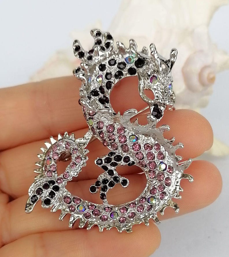 Puede incluir: Broche de drag&oacute;n plateado con pedrer&iacute;a rosa y negra. El dise&ntilde;o del drag&oacute;n presenta escamas detalladas y un cierre de alfiler. Broche decorativo.