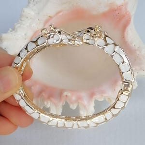 May include: A hinged bangle bracelet featuring two giraffe heads meeting at the center. The bracelet is crafted from gold-toned metal with white enamel patches covering the entire surface, mimicking the natural pattern of a giraffe's coat.