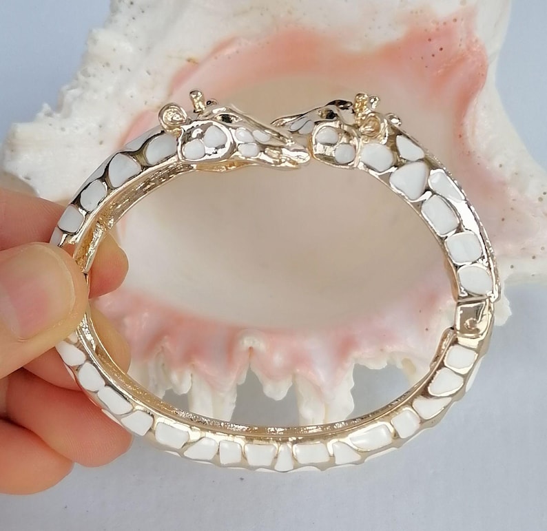 May include: A hinged bangle bracelet featuring two giraffe heads meeting at the center. The bracelet is crafted from gold-toned metal with white enamel patches covering the entire surface, mimicking the natural pattern of a giraffe's coat.