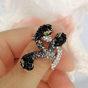 May include: A gold-toned ring featuring two intertwined snakes. The snake bodies are encrusted with black, grey, and clear crystals, creating a gradient effect. Each snake head is detailed with small, vibrant green crystal eyes.