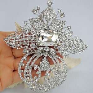 May include: A silver-toned brooch featuring a large rectangular clear crystal at the center, surrounded by intricate filigree metalwork encrusted with numerous small round clear rhinestones and a central teardrop-shaped crystal accent.