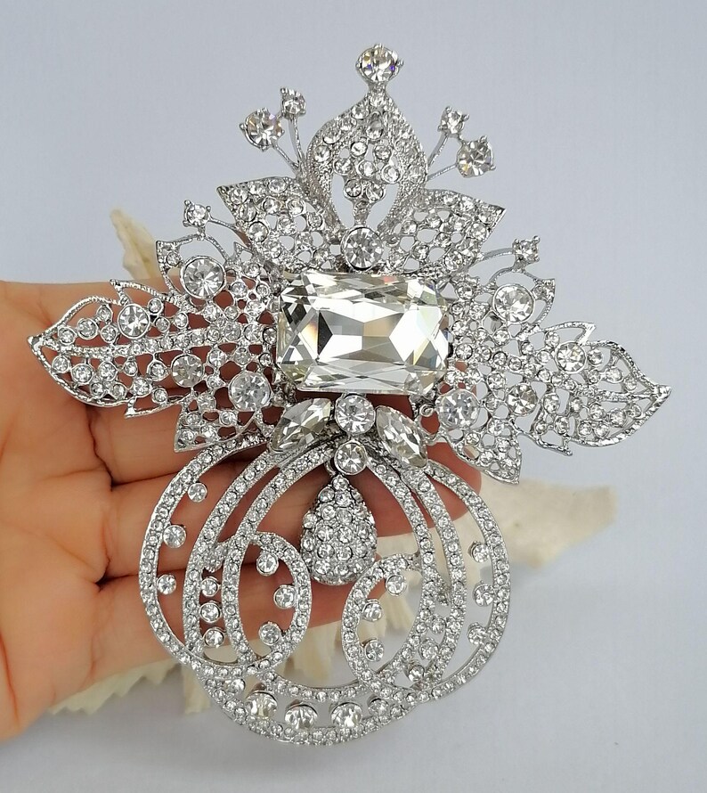 May include: A silver-toned brooch featuring a large rectangular clear crystal at the center, surrounded by intricate filigree metalwork encrusted with numerous small round clear rhinestones and a central teardrop-shaped crystal accent.