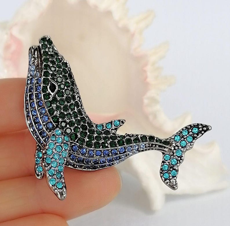 May include: A bejeweled whale brooch in shades of green, blue, and teal. The whale is encrusted with sparkling crystals, creating a shimmering effect. The brooch is a decorative accessory, perfect for adding a touch of marine-inspired elegance to any outfit.