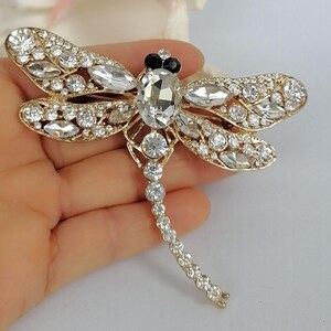 May include: A gold-coloured dragonfly brooch with clear and black crystal accents. The brooch features a large oval crystal body, crystal-studded wings, and a delicate crystal tail. The wings are detailed with smaller crystals.
