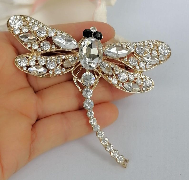 May include: A gold-coloured dragonfly brooch with clear and black crystal accents. The brooch features a large oval crystal body, crystal-studded wings, and a delicate crystal tail. The wings are detailed with smaller crystals.