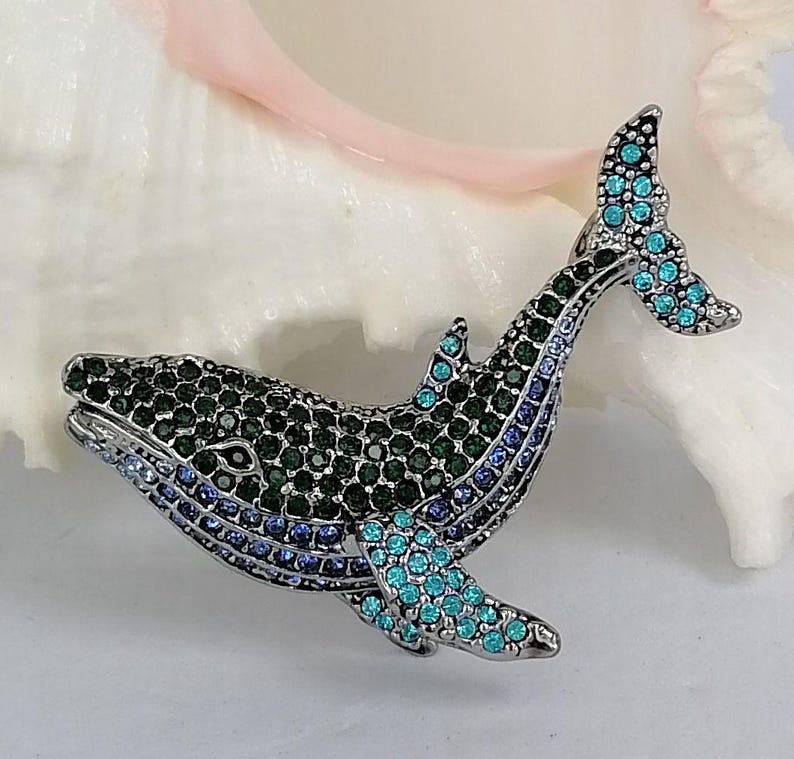 May include: A bejeweled whale brooch with a silver-tone metal finish. The whale is encrusted with green, blue, and teal rhinestones. The brooch is set against a white background.