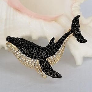 May include: A bejeweled whale brooch. The brooch features a black body with a gold-colored trim and a belly lined with clear crystals. The whale's tail and dorsal fin are also black. The brooch is set against a white background.