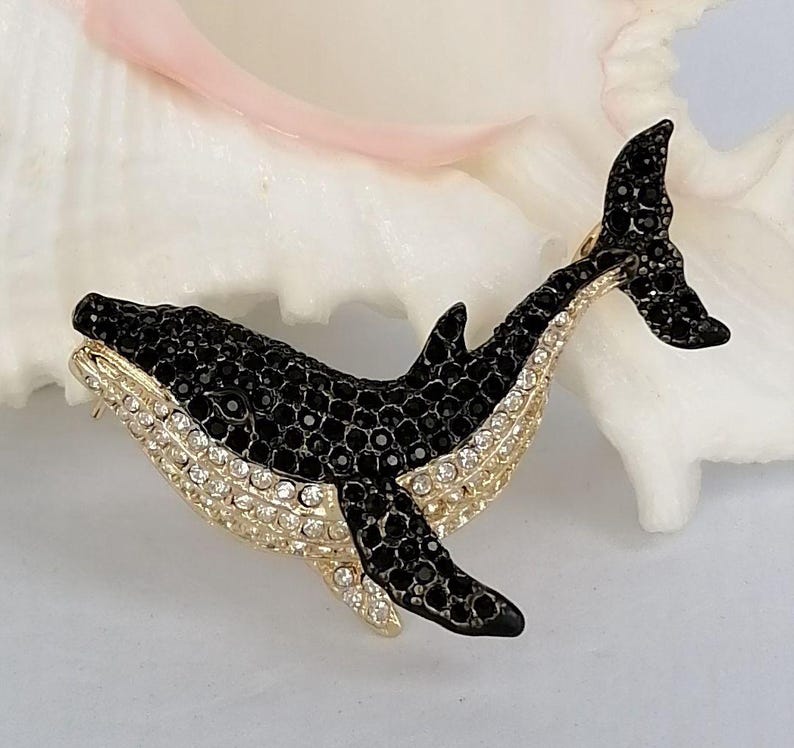 May include: A bejeweled whale brooch. The brooch features a black body with a gold-colored trim and a belly lined with clear crystals. The whale's tail and dorsal fin are also black. The brooch is set against a white background.