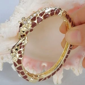 May include: A gold-tone hinged bangle bracelet featuring a giraffe motif with brown enamel spots. The bracelet is accented with a row of clear crystals along the top edge and two giraffe heads meeting at the clasp, finished with a polished metal.