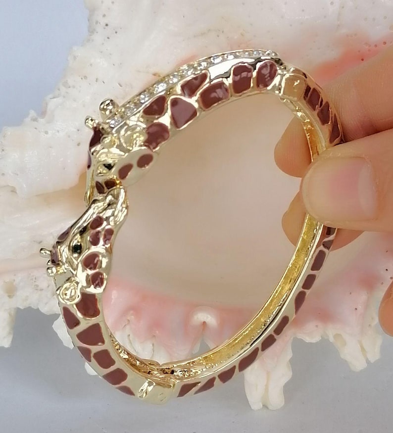May include: A gold-tone hinged bangle bracelet featuring a giraffe motif with brown enamel spots. The bracelet is accented with a row of clear crystals along the top edge and two giraffe heads meeting at the clasp, finished with a polished metal.