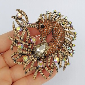 May include: A detailed horse brooch with a brown body and a gold-colored mane and accents. The brooch is adorned with numerous iridescent rhinestones and a large, clear teardrop-shaped stone. The design is intricate and eye-catching, suitable for adding a touch of elegance.