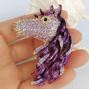 K&ouml;nnte beinhalten: Pferdekopf-Brosche mit goldfarbener Schnauze und lavendelfarbenen Kristallakzenten. Die M&auml;hne des Pferdes ist tiefviolett mit Kristallverzierungen. Die Brosche ist ein dekoratives Accessoire.