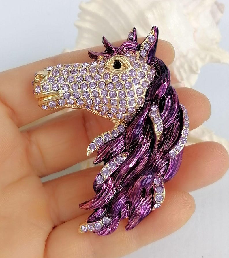 K&ouml;nnte beinhalten: Pferdekopf-Brosche mit goldfarbener Schnauze und lavendelfarbenen Kristallakzenten. Die M&auml;hne des Pferdes ist tiefviolett mit Kristallverzierungen. Die Brosche ist ein dekoratives Accessoire.