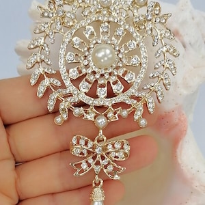 May include: A gold-tone brooch featuring a circular frame adorned with clear rhinestones and small white pearls. The design includes leaf-like branches, a central pearl, and a dangling bow element with a single teardrop-shaped pearl pendant.