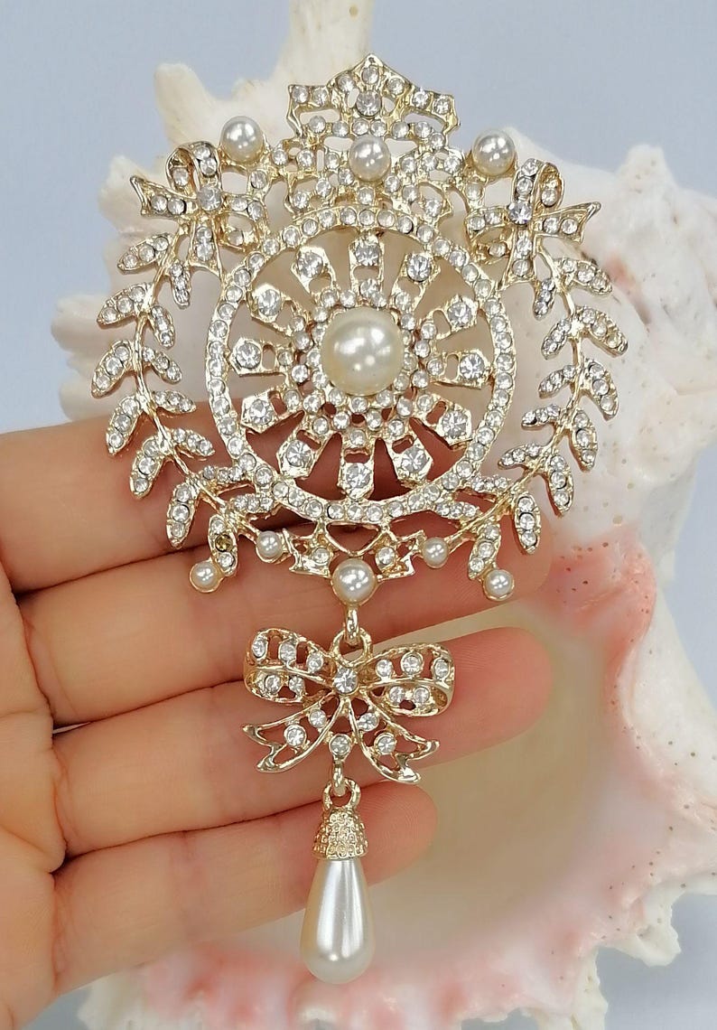 May include: A gold-tone brooch featuring a circular frame adorned with clear rhinestones and small white pearls. The design includes leaf-like branches, a central pearl, and a dangling bow element with a single teardrop-shaped pearl pendant.