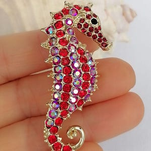 May include: A gold-toned seahorse brooch encrusted with red and iridescent crystals. The seahorse has a detailed design with a curled tail and a black eye. The brooch is a decorative accessory.