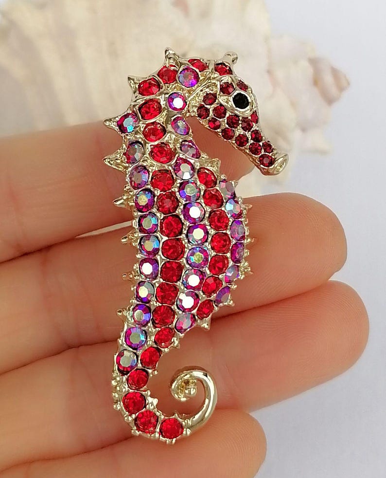 May include: A gold-toned seahorse brooch encrusted with red and iridescent crystals. The seahorse has a detailed design with a curled tail and a black eye. The brooch is a decorative accessory.