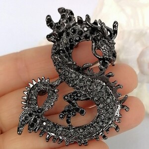 Puede incluir: Broche de drag&oacute;n negro, incrustado con peque&ntilde;as gemas oscuras. El drag&oacute;n est&aacute; en una pose curvada, con escamas detalladas y una expresi&oacute;n feroz. Un accesorio decorativo.