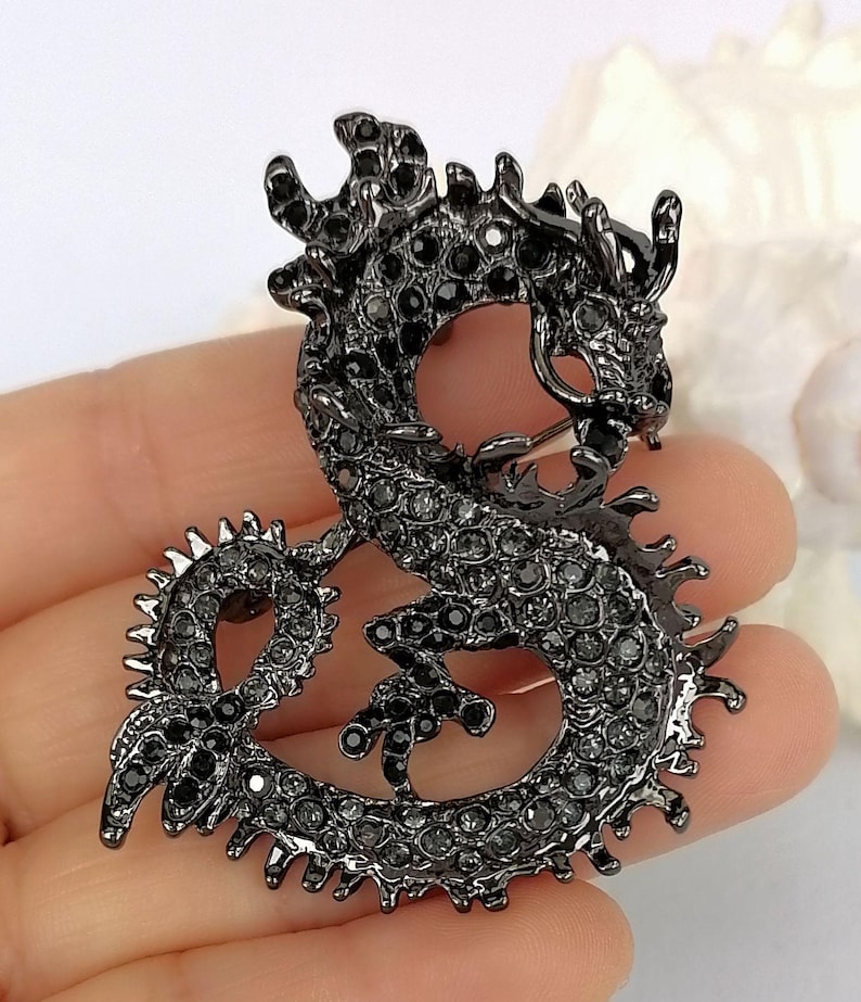 Puede incluir: Broche de drag&oacute;n negro, incrustado con peque&ntilde;as gemas oscuras. El drag&oacute;n est&aacute; en una pose curvada, con escamas detalladas y una expresi&oacute;n feroz. Un accesorio decorativo.