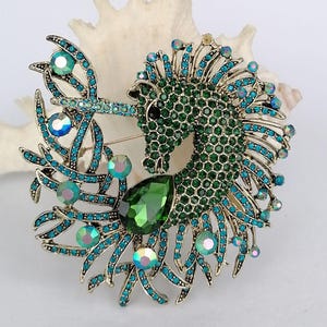 May include: A detailed brooch featuring a seahorse design. The seahorse is primarily green with a textured surface, accented with teal and iridescent crystals. A large, teardrop-shaped green gem is at the base. The brooch has a gold-tone metal finish.