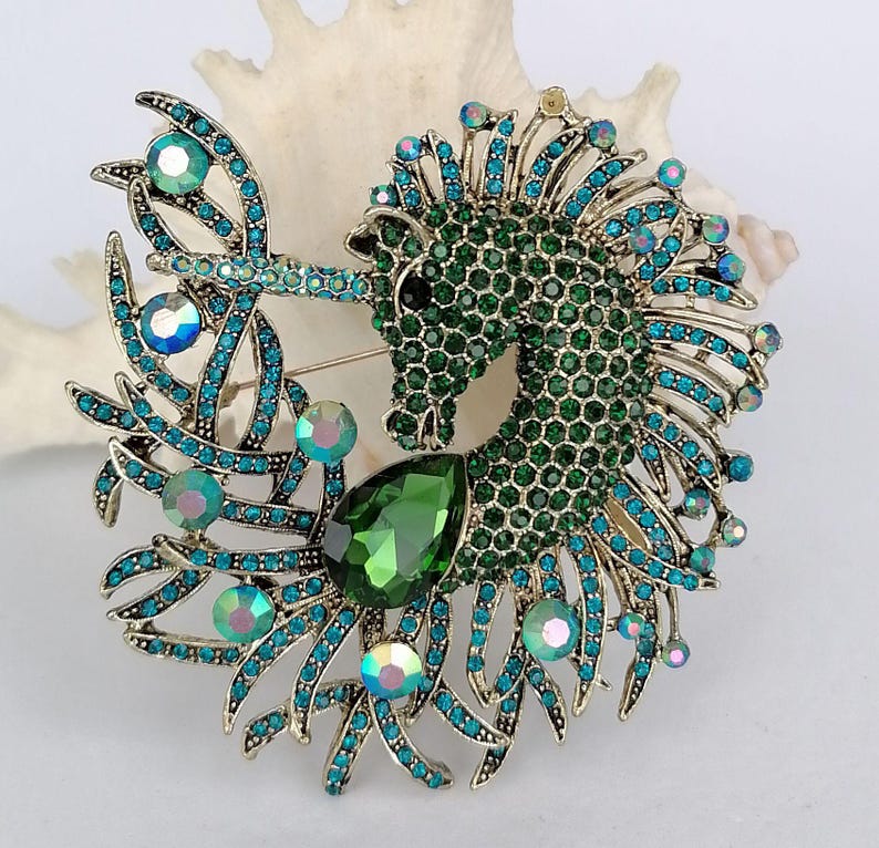 May include: A detailed brooch featuring a seahorse design. The seahorse is primarily green with a textured surface, accented with teal and iridescent crystals. A large, teardrop-shaped green gem is at the base. The brooch has a gold-tone metal finish.