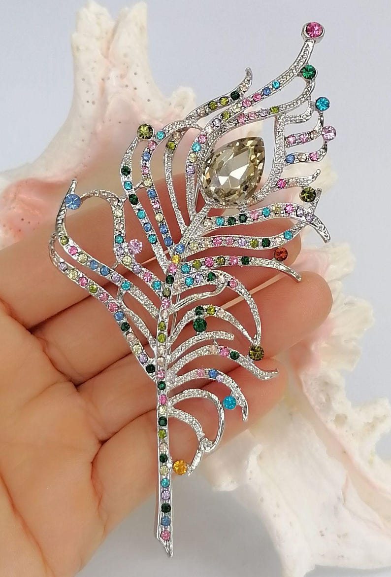 May include: A silver feather brooch, intricately detailed with a variety of colourful gemstones. The brooch features a large, teardrop-shaped central stone and is embellished with smaller gems in pink, green, blue, and yellow.