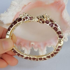 May include: A gold-tone hinged bangle bracelet featuring two giraffe heads meeting at the center. The bracelet is decorated with brown enamel spots across the entire band, mimicking a giraffe's natural coat pattern with a polished finish.