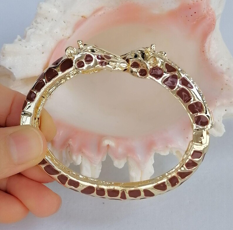 May include: A gold-tone hinged bangle bracelet featuring two giraffe heads meeting at the center. The bracelet is decorated with brown enamel spots across the entire band, mimicking a giraffe's natural coat pattern with a polished finish.