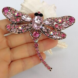 May include: A dragonfly brooch with pink and iridescent crystals. The brooch features a large oval crystal at the centre, with smaller crystals forming the wings and body. The metal has a gold finish.