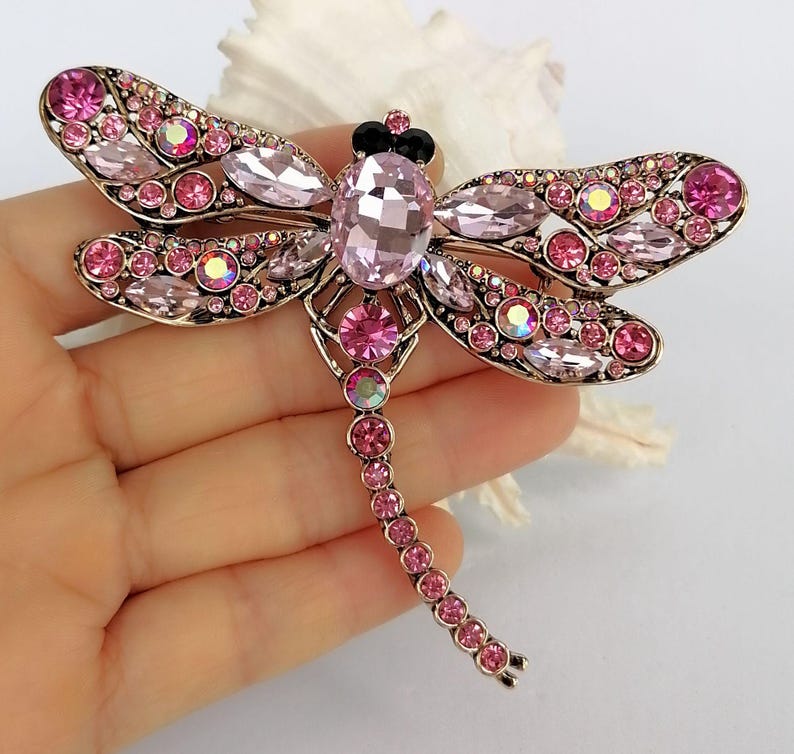 May include: A dragonfly brooch with pink and iridescent crystals. The brooch features a large oval crystal at the centre, with smaller crystals forming the wings and body. The metal has a gold finish.
