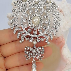May include: A silver-toned brooch featuring a circular frame adorned with clear rhinestones and small white pearls. The design includes a central pearl, leaf-like accents, and a dangling bow charm that supports a single, teardrop-shaped white pearl.