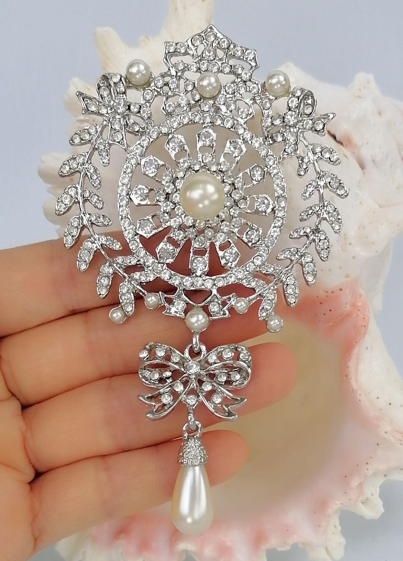 May include: A silver-toned brooch featuring a circular frame adorned with clear rhinestones and small white pearls. The design includes a central pearl, leaf-like accents, and a dangling bow charm that supports a single, teardrop-shaped white pearl.