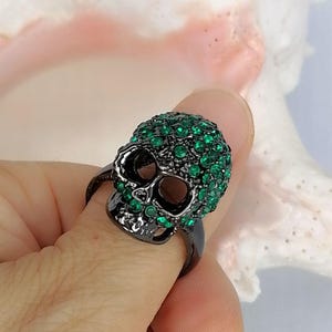 Op de afbeelding: Een zwarte metalen ring in de vorm van een schedel, met een donkere afwerking en bedekt met talloze kleine, levendige groene strass-steentjes die het gehele oppervlak van het schedelontwerp bedekken, inclusief de voorhoofd- en jukbeenderen.
