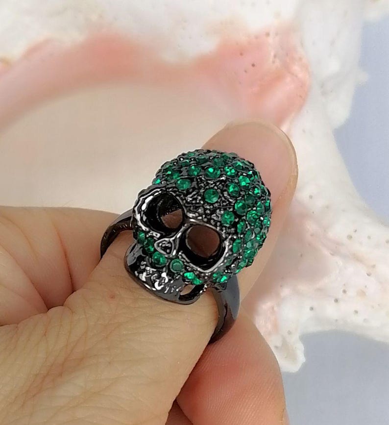 Op de afbeelding: Een zwarte metalen ring in de vorm van een schedel, met een donkere afwerking en bedekt met talloze kleine, levendige groene strass-steentjes die het gehele oppervlak van het schedelontwerp bedekken, inclusief de voorhoofd- en jukbeenderen.