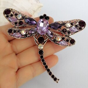 May include: A dragonfly brooch with a gold-tone metal frame. The brooch is decorated with various purple and clear crystals. The dragonfly's body features a large, oval lavender crystal, and its wings are embellished with smaller crystals in shades of purple and clear.