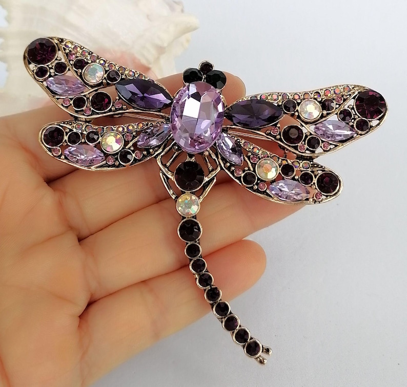 May include: A dragonfly brooch with a gold-tone metal frame. The brooch is decorated with various purple and clear crystals. The dragonfly's body features a large, oval lavender crystal, and its wings are embellished with smaller crystals in shades of purple and clear.