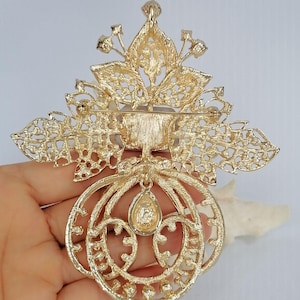 May include: A gold-tone metal brooch featuring an intricate filigree leaf design at the top and a circular frame at the bottom. A single teardrop-shaped stone hangs from the center of the circular frame, and a horizontal pin clasp is on the back.