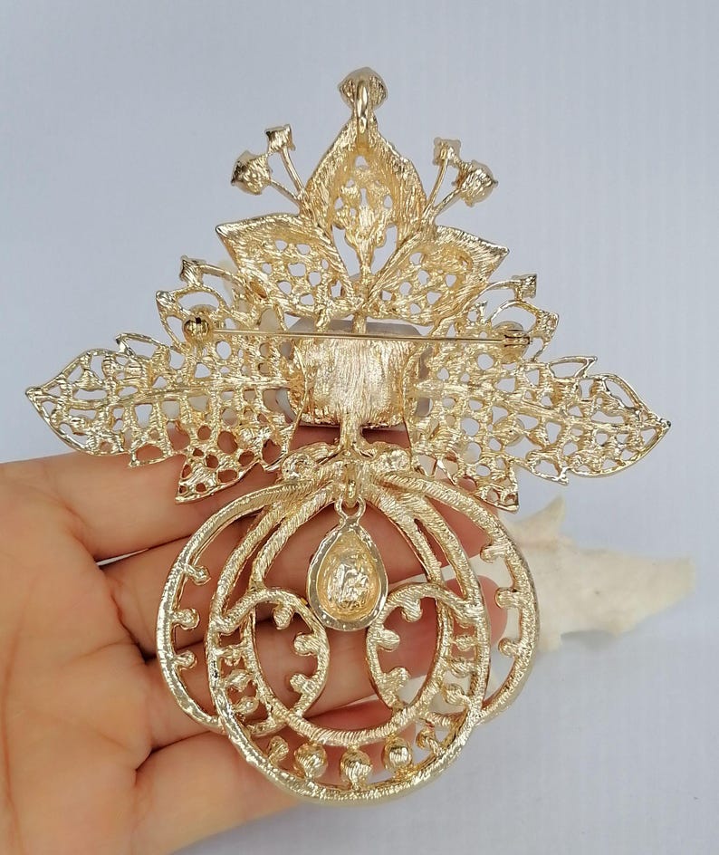 May include: A gold-tone metal brooch featuring an intricate filigree leaf design at the top and a circular frame at the bottom. A single teardrop-shaped stone hangs from the center of the circular frame, and a horizontal pin clasp is on the back.