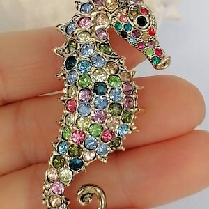 May include: A gold-toned seahorse brooch encrusted with colorful rhinestones. The brooch features a detailed design with a curled tail and a textured body. The rhinestones are in various colors, including pink, blue, green, and yellow.
