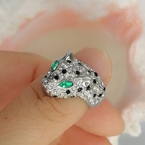 May include: A silver-toned ring shaped like a panther head, featuring a pav&eacute; setting of clear white stones, scattered black stones for spots, and two marquise-cut green stones serving as the eyes of the feline figure.