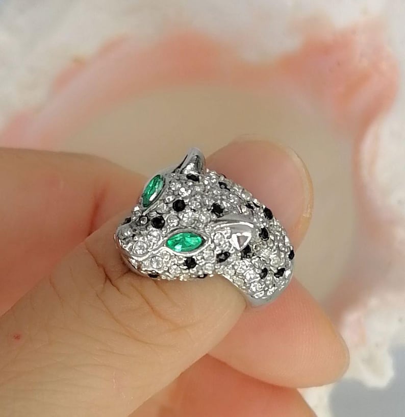 May include: A silver-toned ring shaped like a panther head, featuring a pav&eacute; setting of clear white stones, scattered black stones for spots, and two marquise-cut green stones serving as the eyes of the feline figure.
