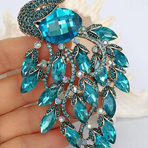 Op de afbeelding: Een pauwbroche met een blauwgroen en blauw kleurenschema. De broche heeft een grote, gefacetteerde blauwe edelsteen in het midden, omringd door kleinere blauwe en heldere kristallen. De staart van de pauw bestaat uit druppelvormige blauwe kristallen.