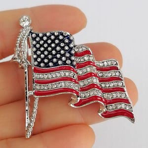 Pode incluir: Broche patriótico em forma de bandeira americana. A bandeira apresenta listras vermelhas e brancas, um campo azul com estrelas brancas e é adornada com strass brilhantes. O broche tem um acabamento em metal prateado.