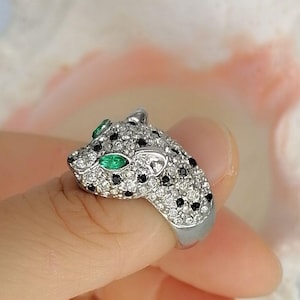 May include: A silver-toned ring shaped like a panther head, featuring a pav&eacute; setting of clear white crystals and small black stones. The panther's eyes are set with marquise-cut green stones, and the band is a smooth, polished silver-toned metal.