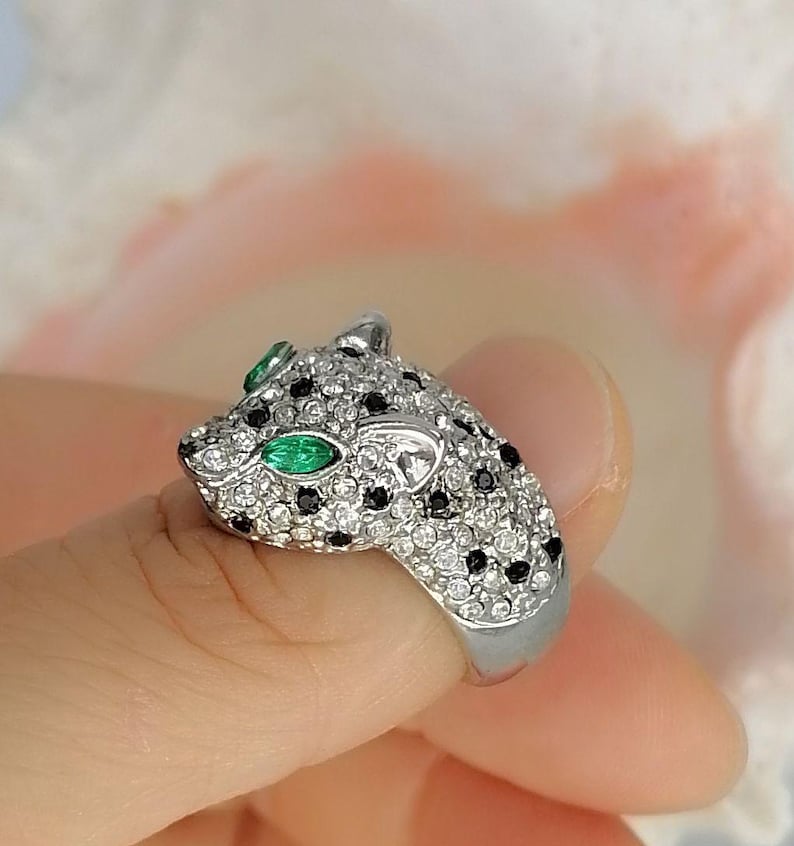 May include: A silver-toned ring shaped like a panther head, featuring a pav&eacute; setting of clear white crystals and small black stones. The panther's eyes are set with marquise-cut green stones, and the band is a smooth, polished silver-toned metal.