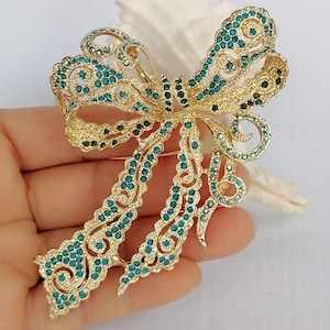 May include: A gold-toned bow brooch with intricate filigree detailing. The bow is embellished with teal and dark green rhinestones, creating a sparkling effect. The brooch is held in a hand.