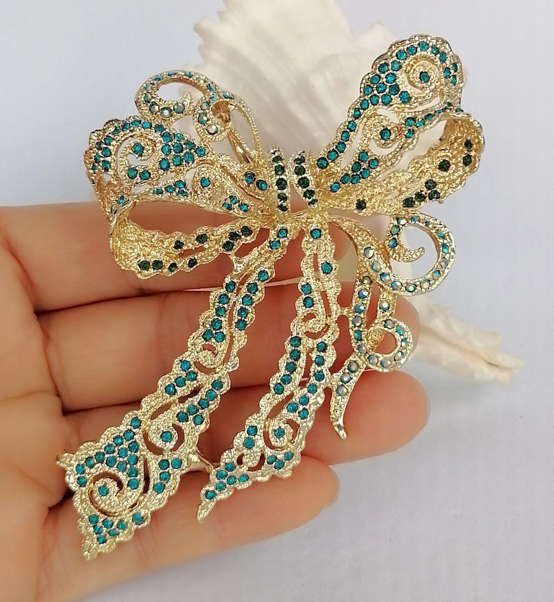 May include: A gold-toned bow brooch with intricate filigree detailing. The bow is embellished with teal and dark green rhinestones, creating a sparkling effect. The brooch is held in a hand.