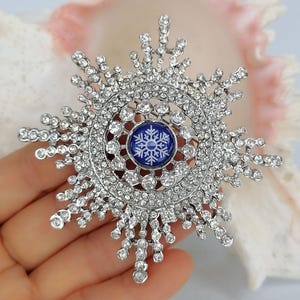 K&ouml;nnte beinhalten: Eine silberfarbene Schneeflocken-Brosche mit zahlreichen klaren, funkelnden Strasssteinen in radialer Anordnung. In der Mitte der Brosche befindet sich ein kreisf&ouml;rmiger blauer Cabochon mit einer wei&szlig;en Schneeflockengrafik auf der Oberfl&auml;che.
