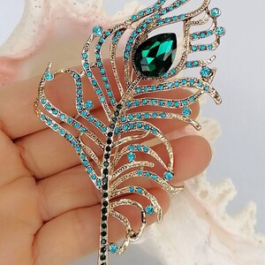 May include: Gold-coloured metal peacock feather brooch with teal and black accents. The brooch features teal-coloured rhinestones and a large emerald-green teardrop-shaped stone. A decorative accessory.